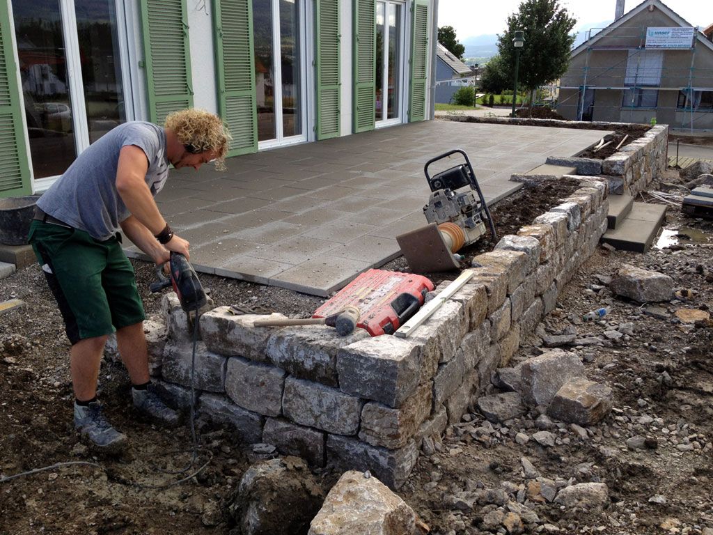 Ein Arbeiter bearbeitet mit einem Bohrhammer eine Steinmauer auf einer geschäftigen Baustelle.