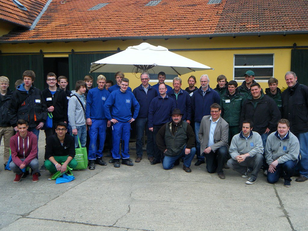 Gruppe posiert vor gelbem Gebäude; einige in blauer Arbeitskleidung, andere in Freizeitkleidung.