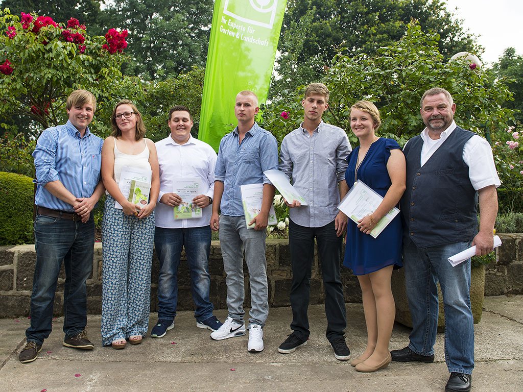 Sieben Personen posieren lächelnd in einem Garten, halten Dokumente; feierliche, offizielle Stimmung.