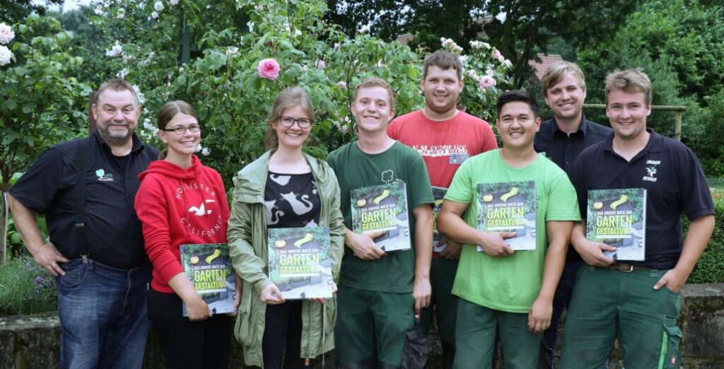Acht Personen lächeln, halten Bücher zur Gartengestaltung; im Hintergrund blühende Rosen und Pflanzen.