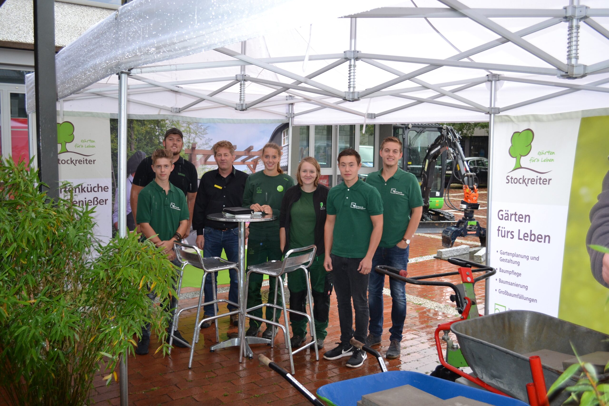 Eine Gruppe in grüner Arbeitskleidung posiert lächelnd unter einem Pavillon bei einer Gartenbau-Veranstaltung.