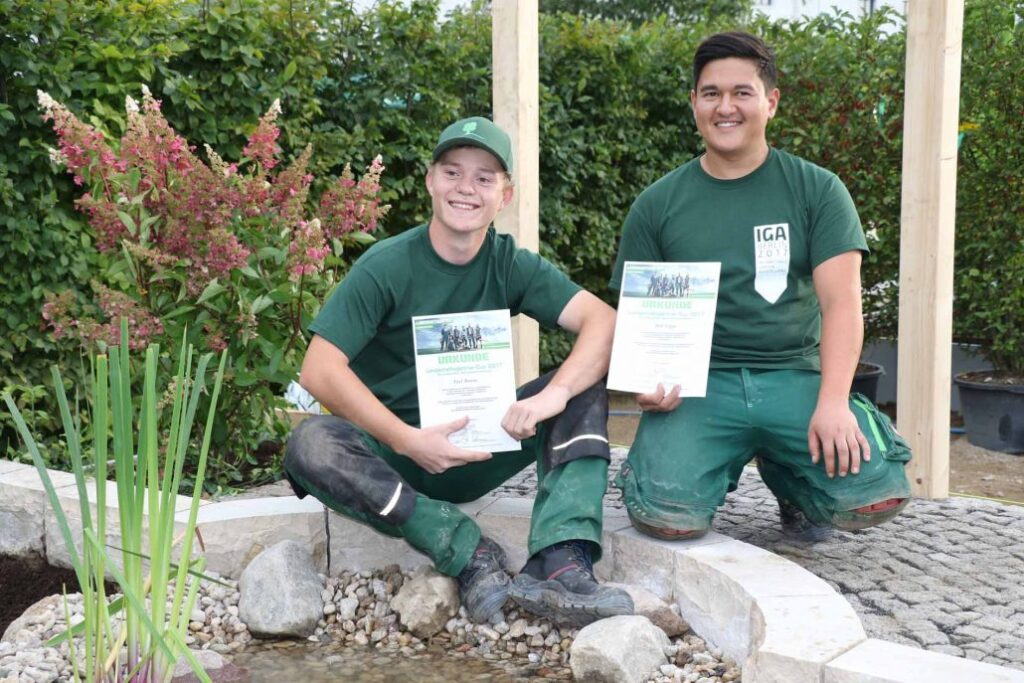 Zwei Gärtner in grüner Arbeitskleidung halten stolz Zertifikate in einem Garten.