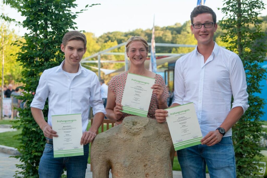 Drei Personen in weißen Hemden halten stolz Dokumente, umgeben von grüner Vegetation und einer Skulptur.