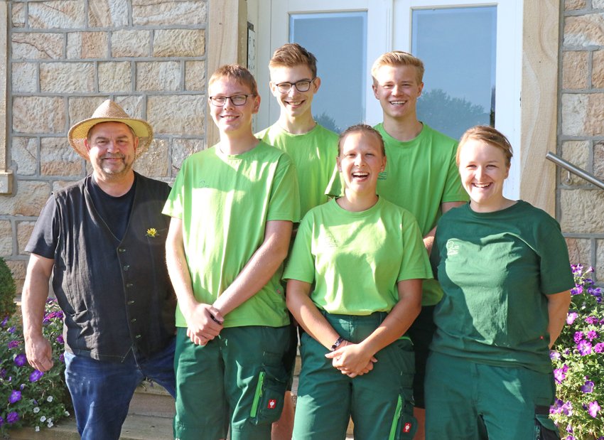 Sechs Personen in grüner Kleidung lächeln vor einer Steinmauer, eine trägt einen Strohhut.