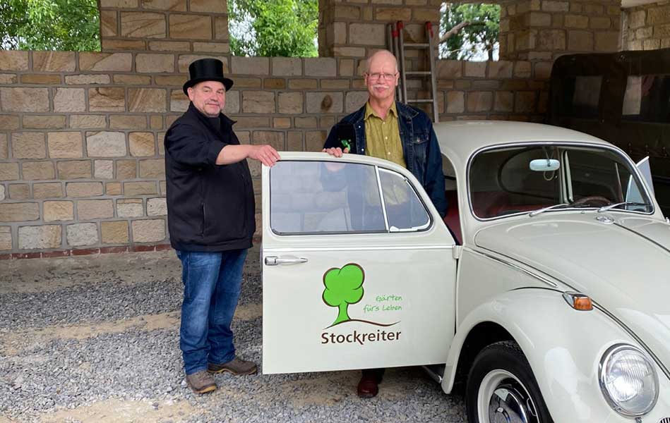 Zwei Männer stehen neben einem cremefarbenen VW Käfer mit "Stockreiter" Logo vor einer Steinmauer.