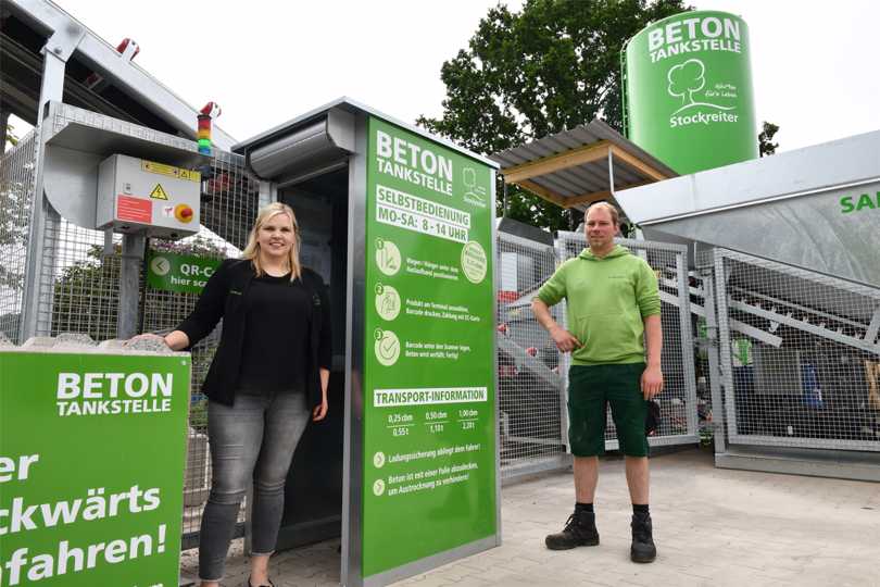 Zwei Personen posieren vor einer Betontankstelle mit grünen Schildern und einem großen Behälter.