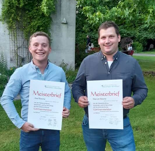 Zwei Personen halten stolz ihre Meisterbriefe vor einer efeubewachsenen Wand und lächeln in die Kamera.