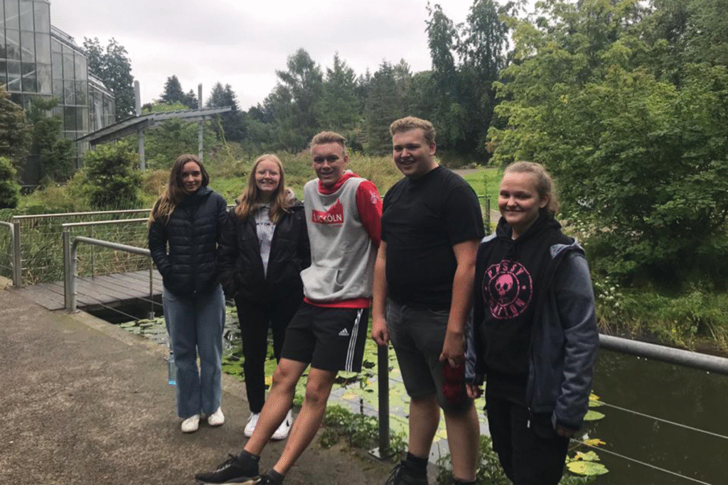 Fünf junge Menschen posieren lächelnd vor einem Teich in einem grünen Park mit Bäumen und Gewächshaus.