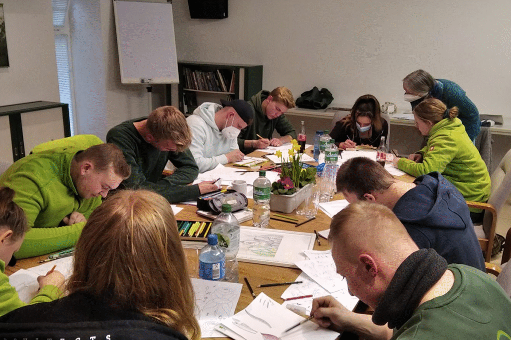 Menschen arbeiten konzentriert an einem Tisch, einige tragen Masken. Eine Person gibt Anweisungen.
