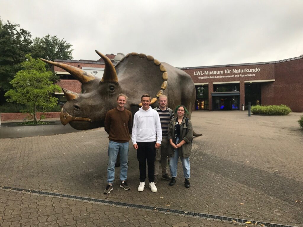 Vier Personen posieren vor einer realistischen Triceratops-Statue vor dem LWL-Museum für Naturkunde.