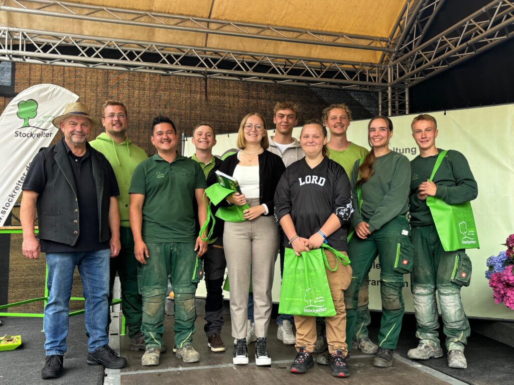 Eine Gruppe von zehn Personen in grüner Arbeitskleidung posiert lächelnd mit "Stockreiter"-Taschen auf einer Bühne.