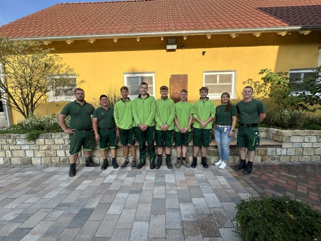 Neun Menschen in grüner Kleidung posieren lächelnd vor einem gelben Gebäude mit rotem Dach.