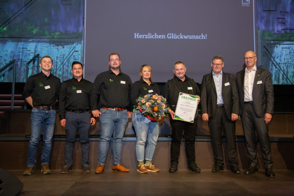 Sieben Personen posieren lächelnd bei einer Preisverleihung. Eine Person hält Blumen, eine andere eine Urkunde.