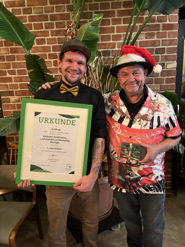 Zwei Männer lächeln, einer hält eine Urkunde, der andere eine CD, beide in festlicher Kleidung.