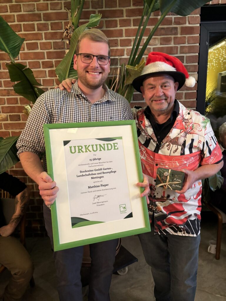 Zwei Männer halten eine Urkunde für 15 Jahre Mitarbeit, einer trägt ein Weihnachtshemd und -hut.