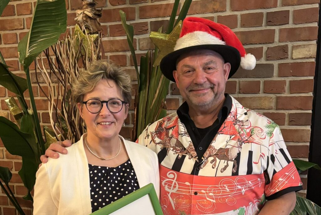 Zwei Personen lächeln; die Frau hält eine Urkunde, der Mann trägt eine Weihnachtsmütze und hält ein Geschenk.