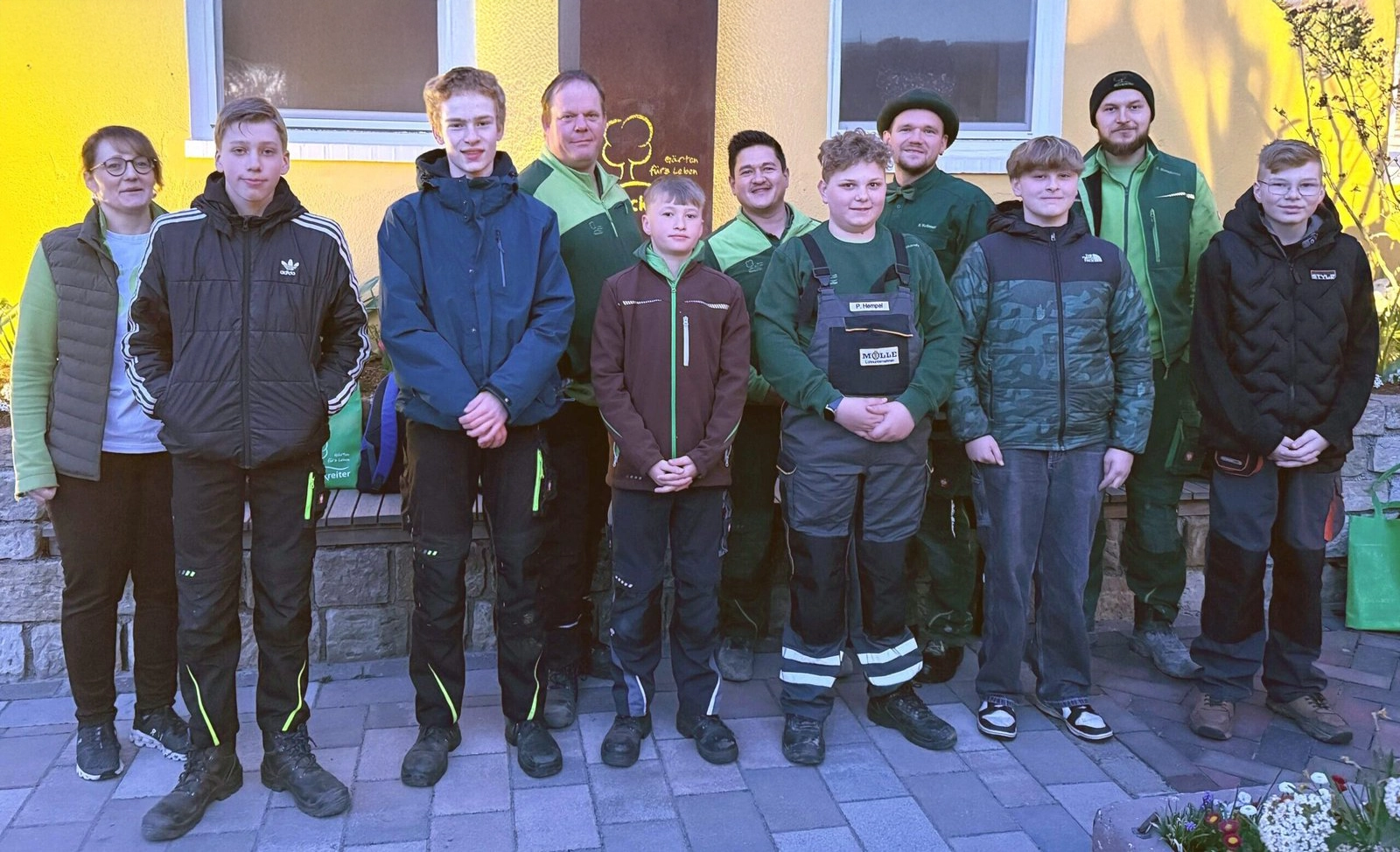 Elf Personen in Outdoor-Kleidung posieren vor einer gelben Wand für ein Gruppenfoto.
