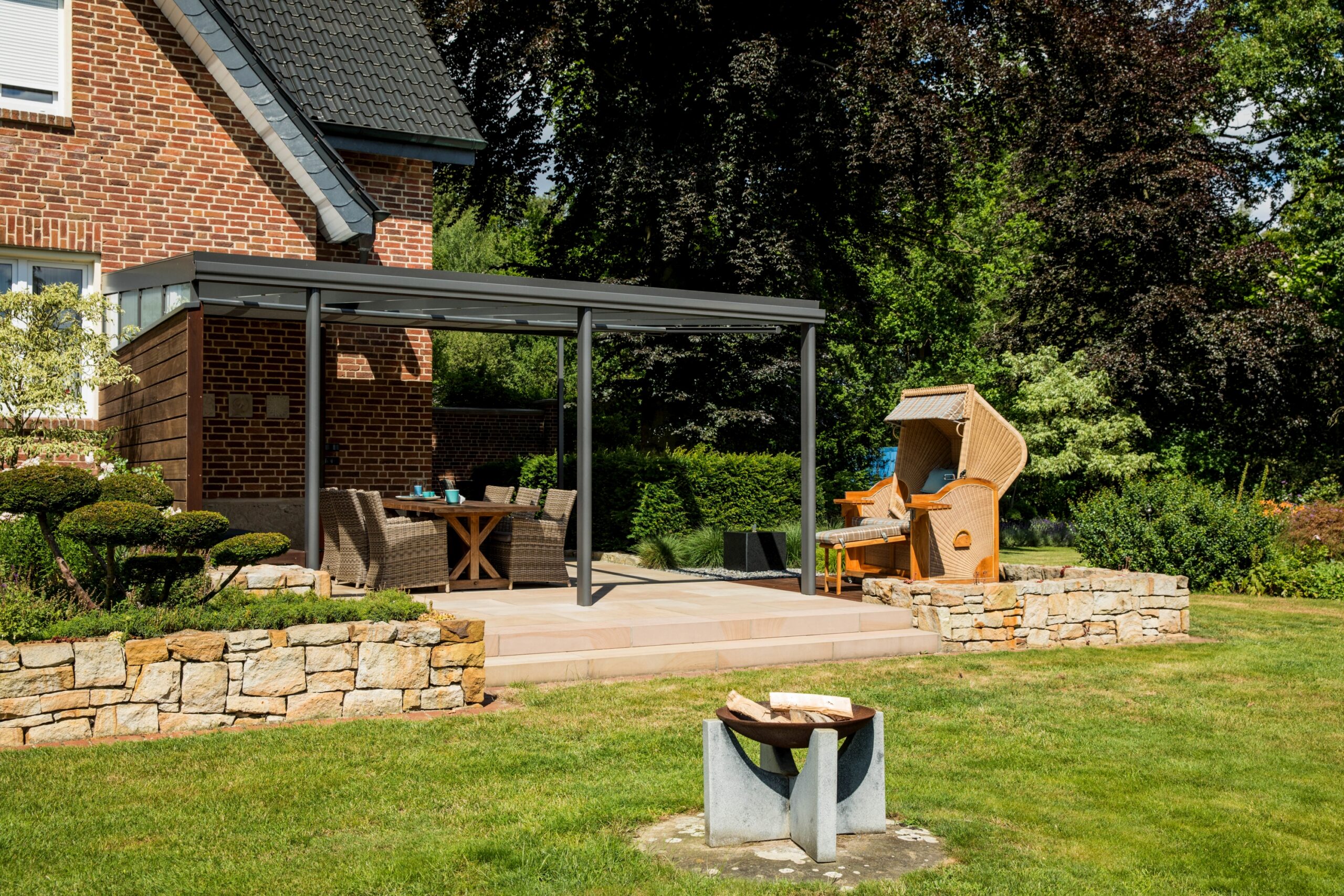 Gepflegter Garten mit Terrassendach, Rattanmöbeln, Strandkorb und Feuerstelle neben Backsteinhaus.
