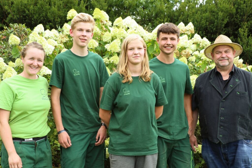 Fünf Personen in grüner Kleidung stehen lächelnd in einem blühenden Garten, eine trägt einen Strohhut.