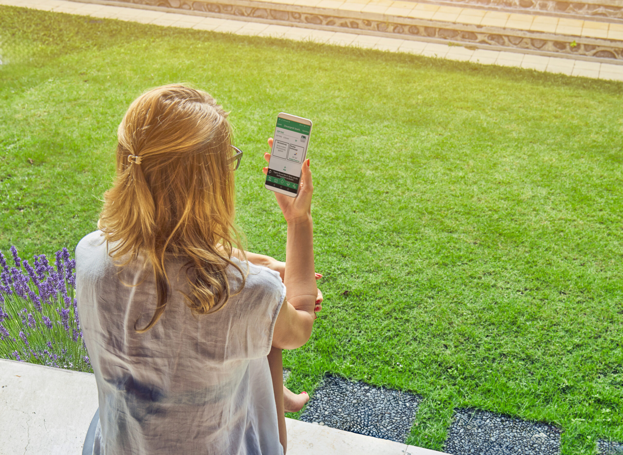 Frau mit Brille sitzt entspannt auf Terrasse und schaut auf ihr Smartphone, umgeben von grünem Rasen.