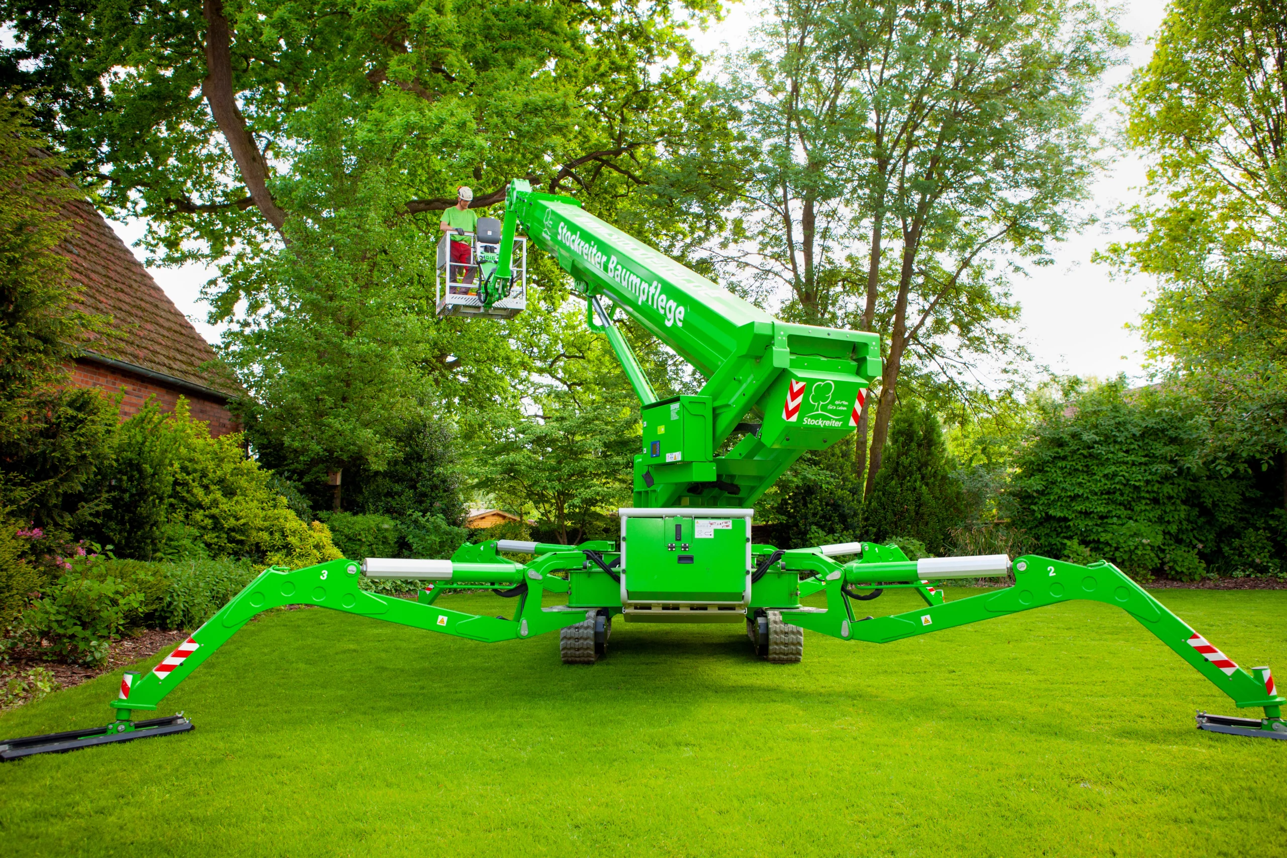 Eine grüne Hebebühne im Garten hebt eine Person zur Baumpflege. Haus und Bäume im Hintergrund.