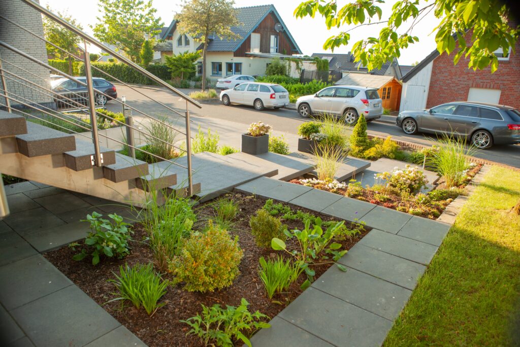 Moderne Gartengestaltung mit Pflanzen und Betonstufen vor einem Wohnhaus, sonniges Wetter.