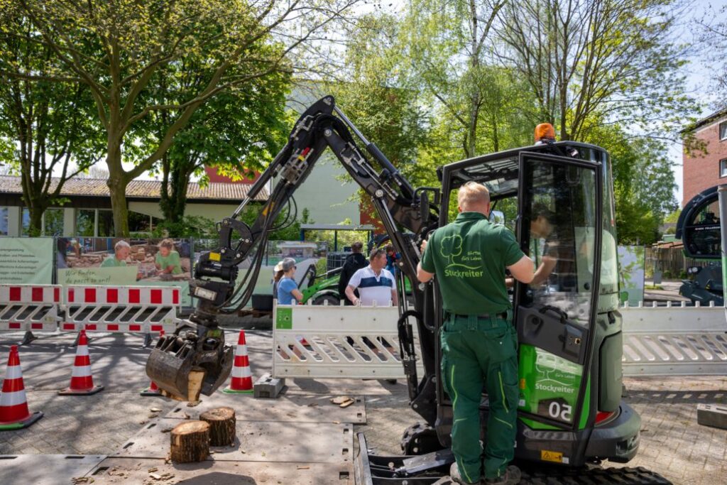 Ein Arbeiter bedient eine grüne Baumaschine neben Baumstämmen auf einer städtischen Baustelle.