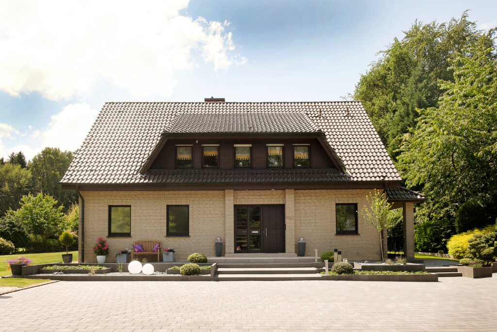 Ein Einfamilienhaus mit hellem Klinker, dunklem Satteldach, umgeben von grüner Landschaft.