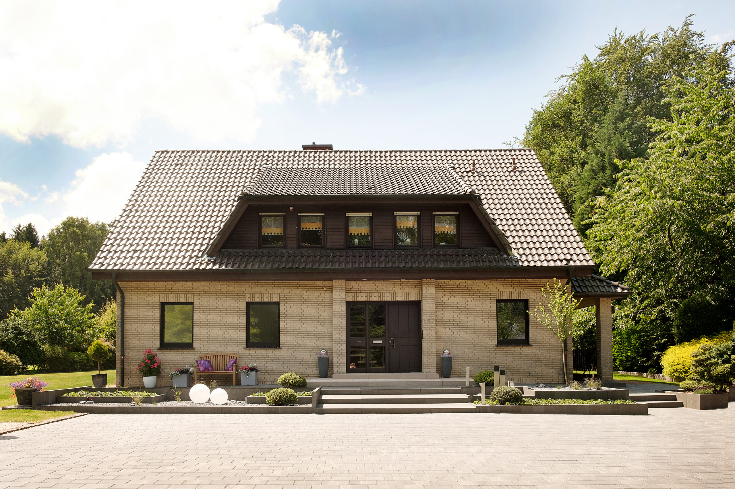 Ein Einfamilienhaus mit hellem Klinker, dunklem Satteldach, umgeben von grüner Landschaft.