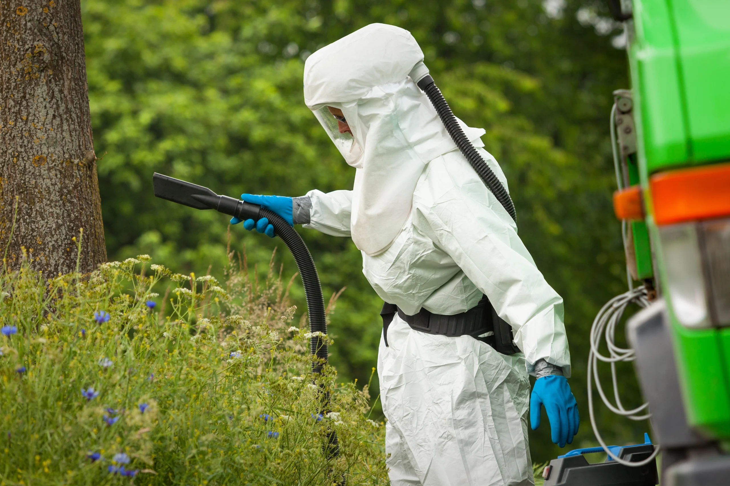 Person im Schutzanzug arbeitet mit Absaugvorrichtung in dichter Vegetation, umgeben von Pflanzen.