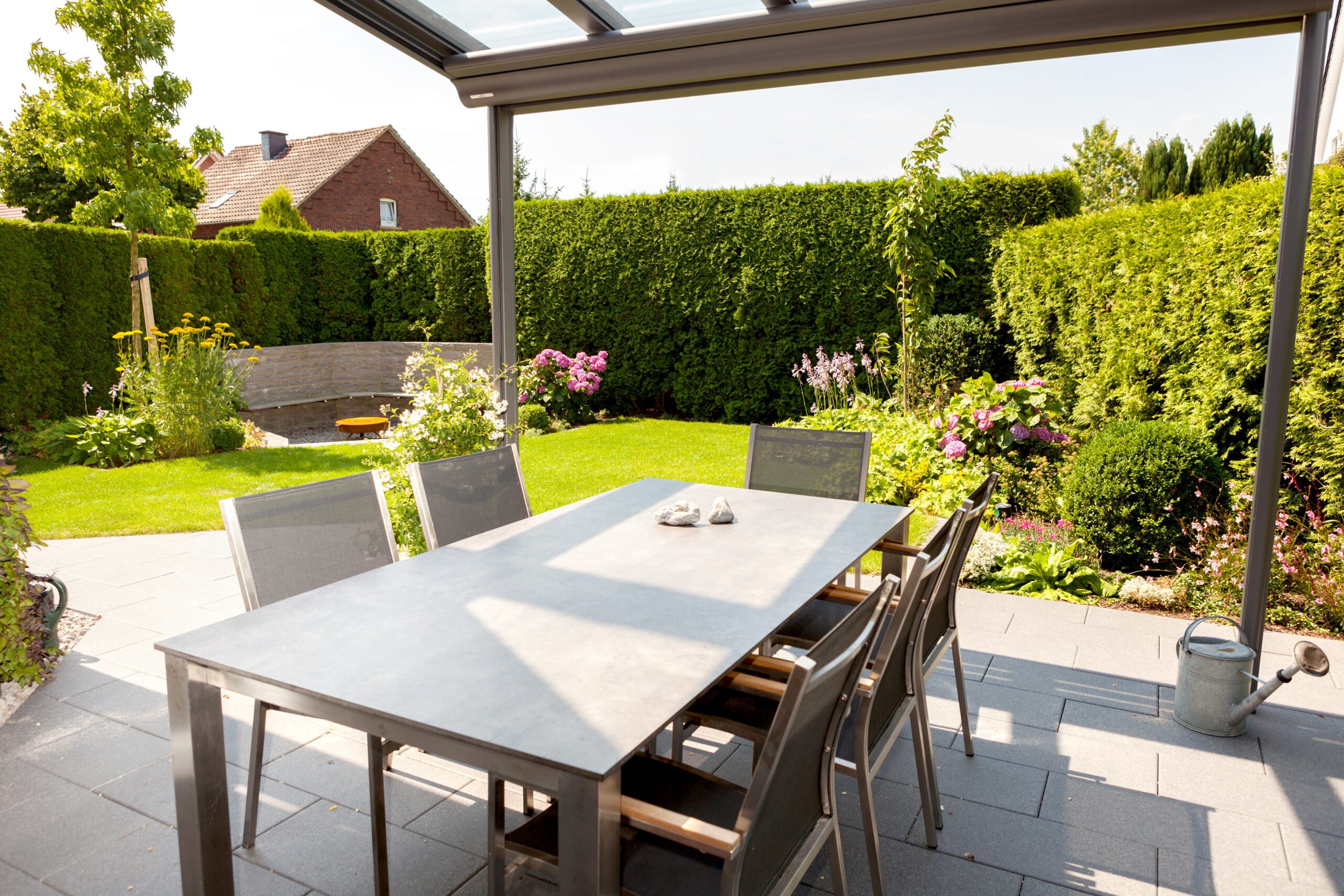 Moderner Terrassenbereich mit Metalltisch, Stühlen, Gießkanne und blühenden Pflanzen im Hintergrund.
