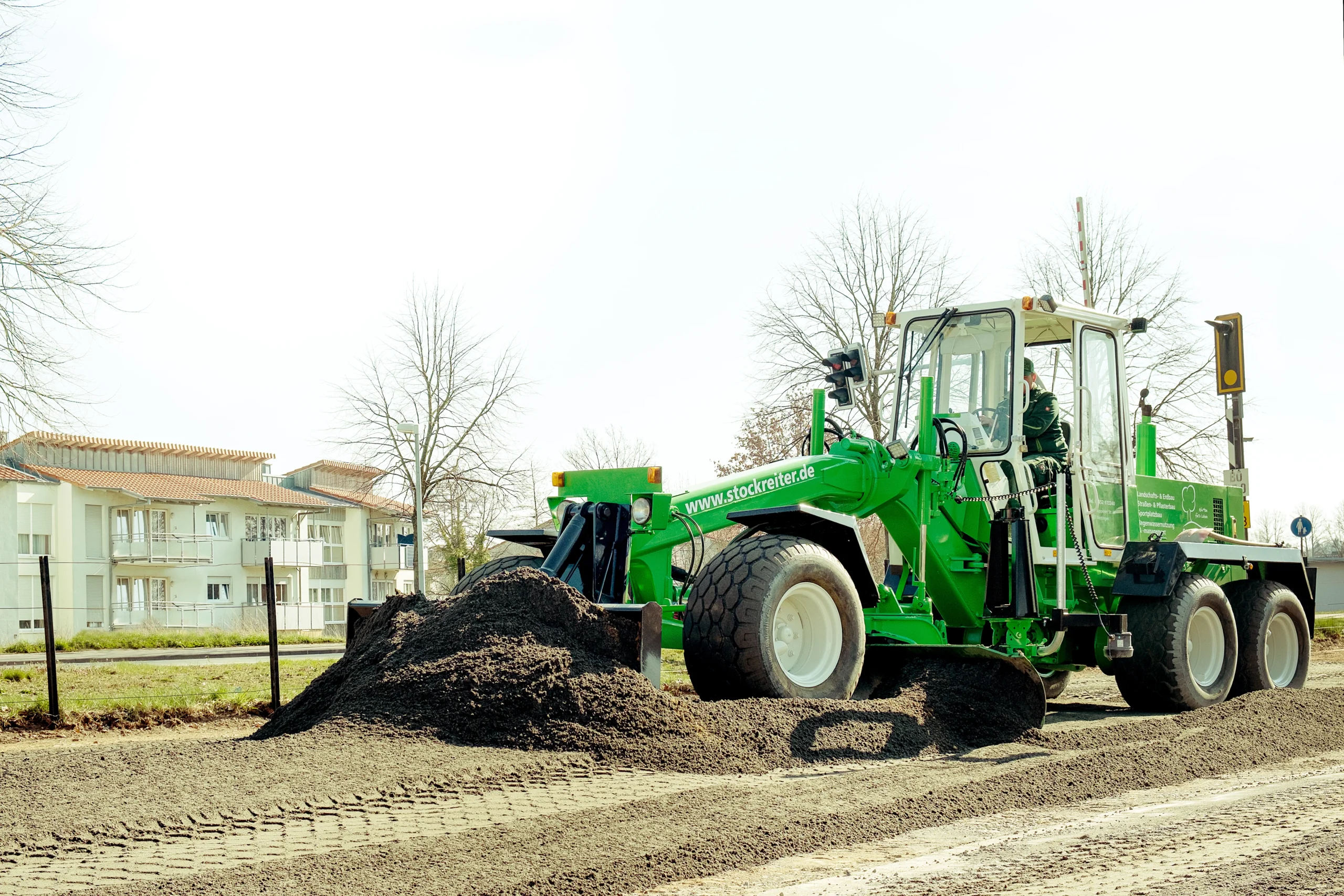 Grüner Grader bewegt Erde auf Baustelle, umgeben von Wohngebäuden und kahlen Bäumen im Hintergrund.