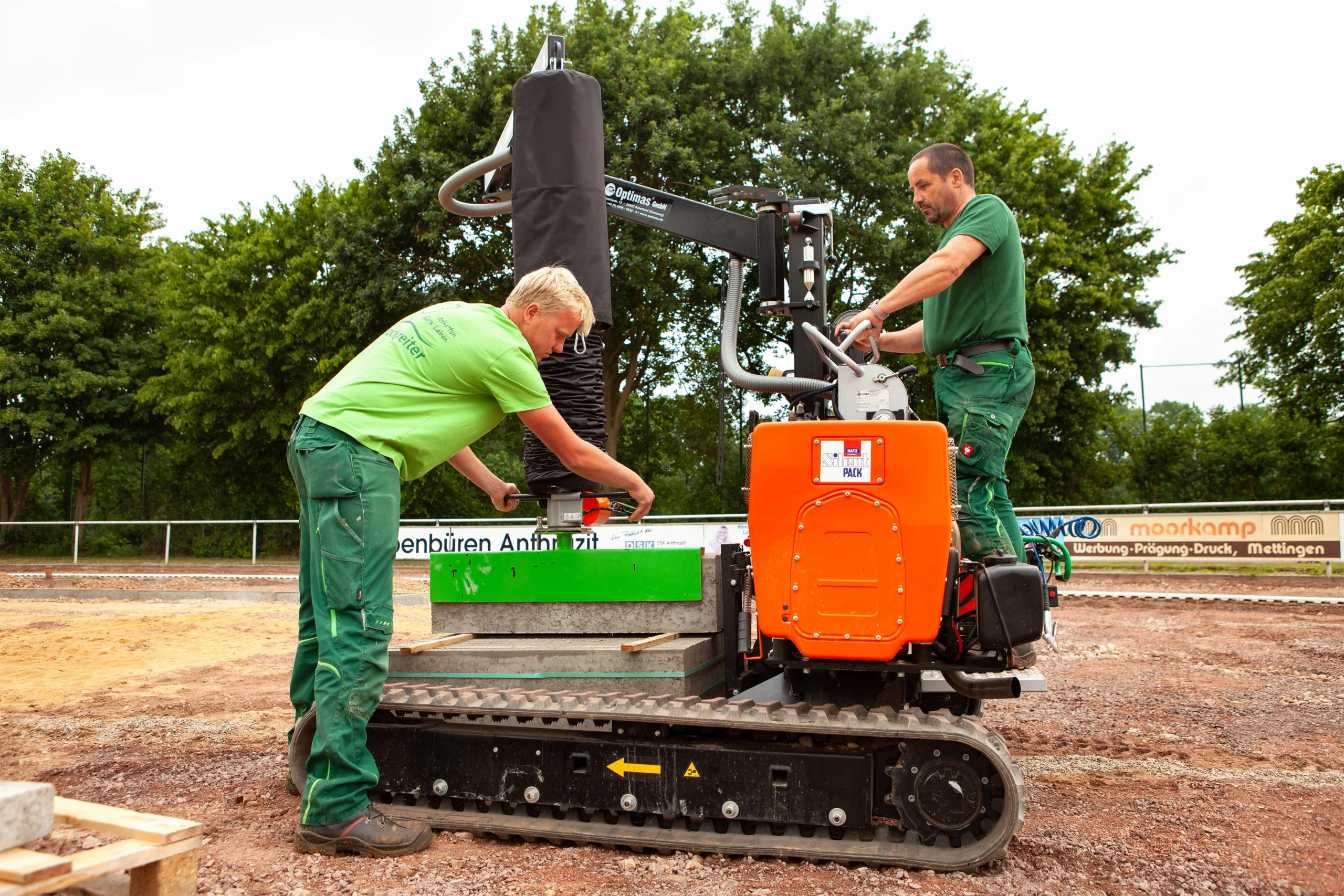 Zwei Arbeiter in grüner Kleidung bedienen eine orangefarbene Maschine, die einen Betonblock bewegt.