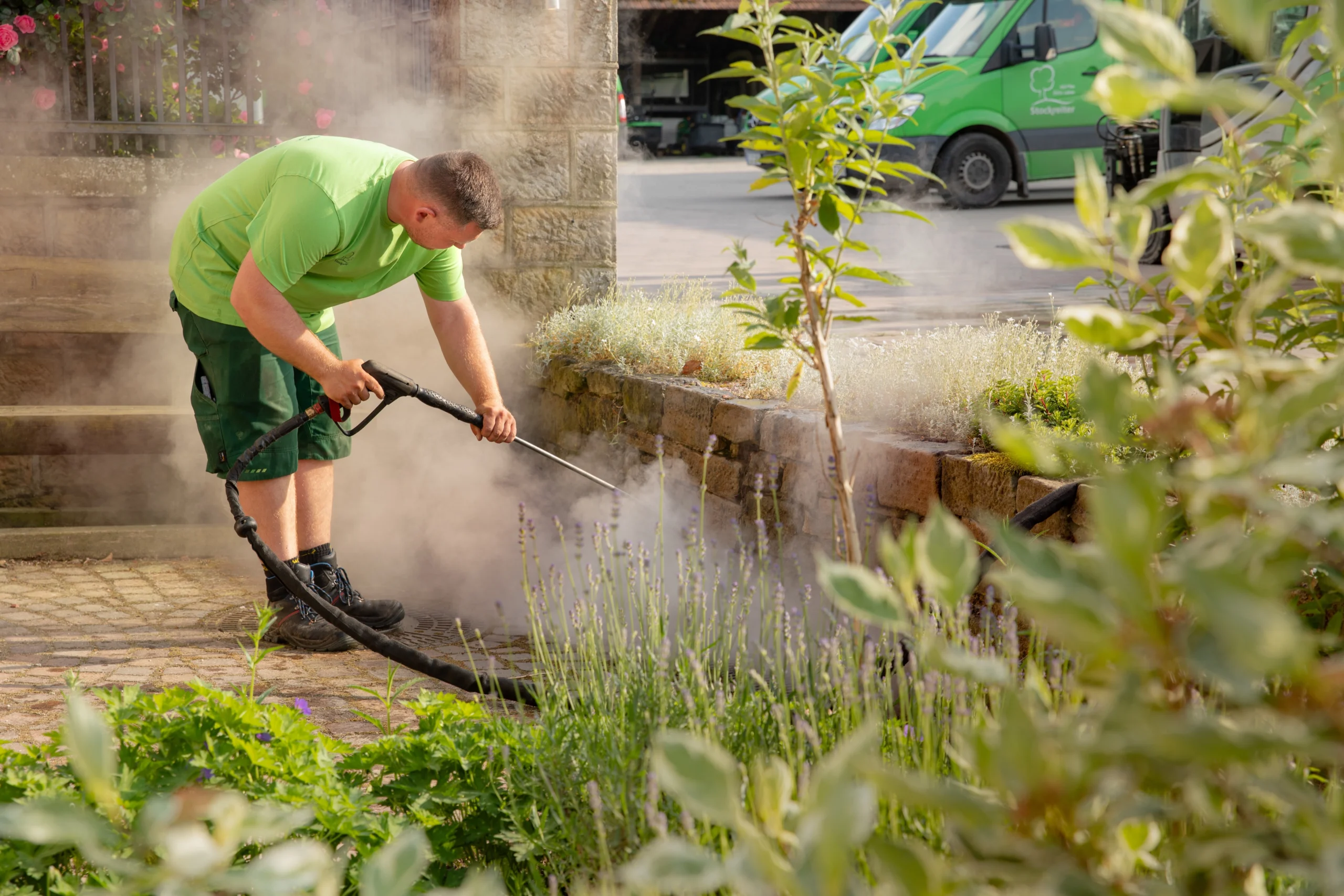 Person reinigt mit Hochdruckreiniger eine gepflasterte Fläche, umgeben von Vegetation und Dampf.