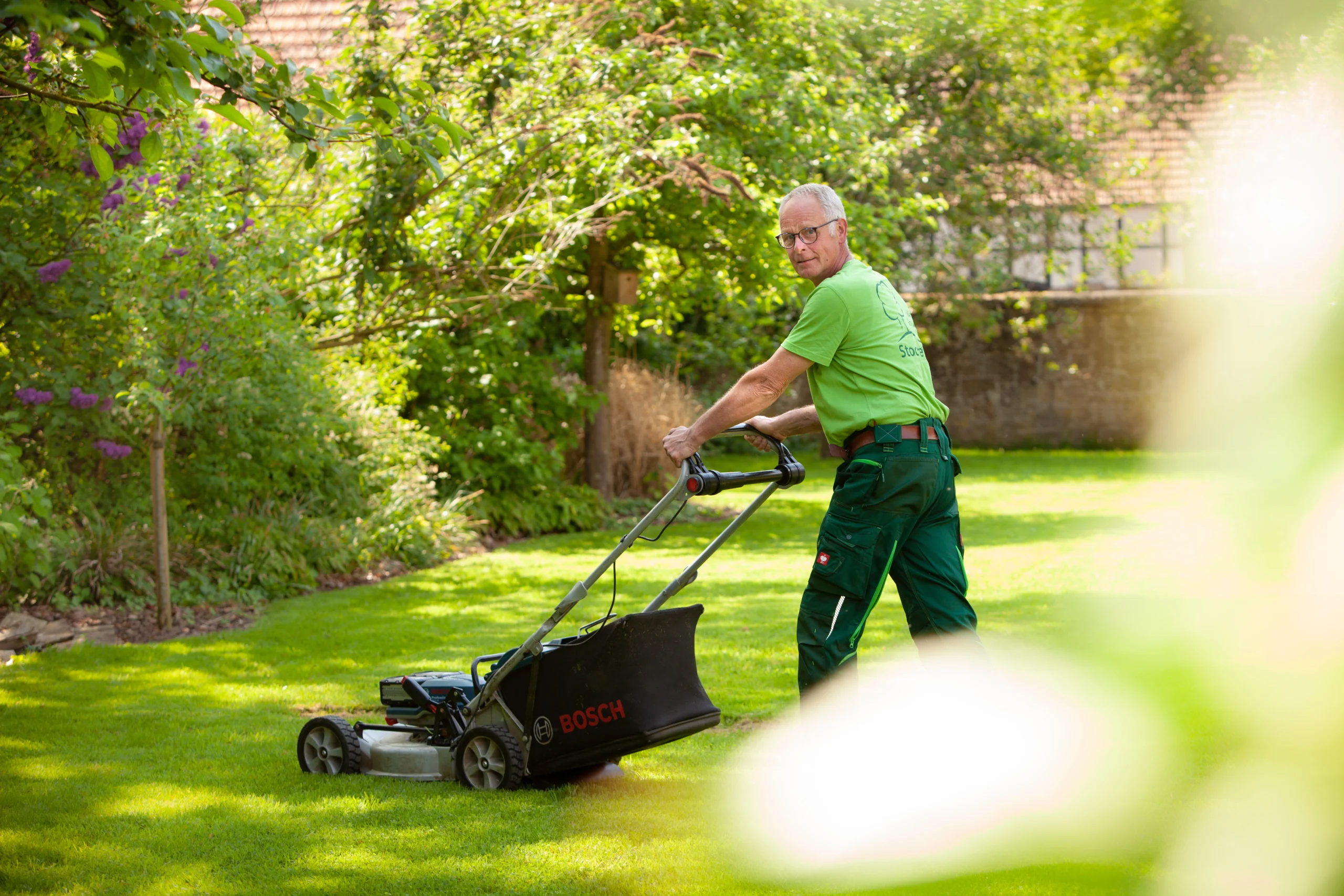 Eine Person in grüner Kleidung mäht mit einem Bosch-Rasenmäher den Rasen in einem grünen Garten.