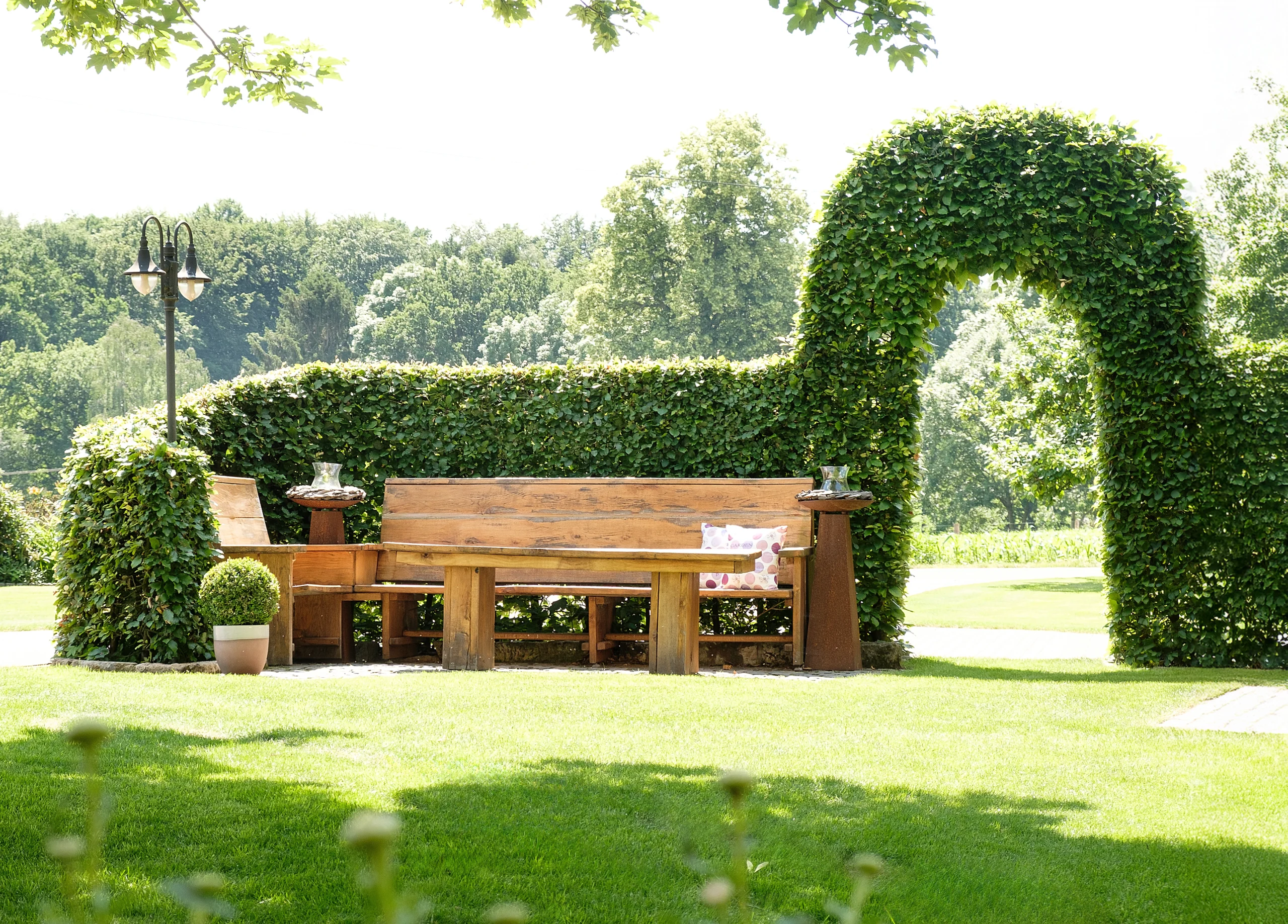 Idyllische Gartenszene mit Holzbank, Tisch, dekorativen Säulen und gepflegter Hecke im Sonnenlicht.