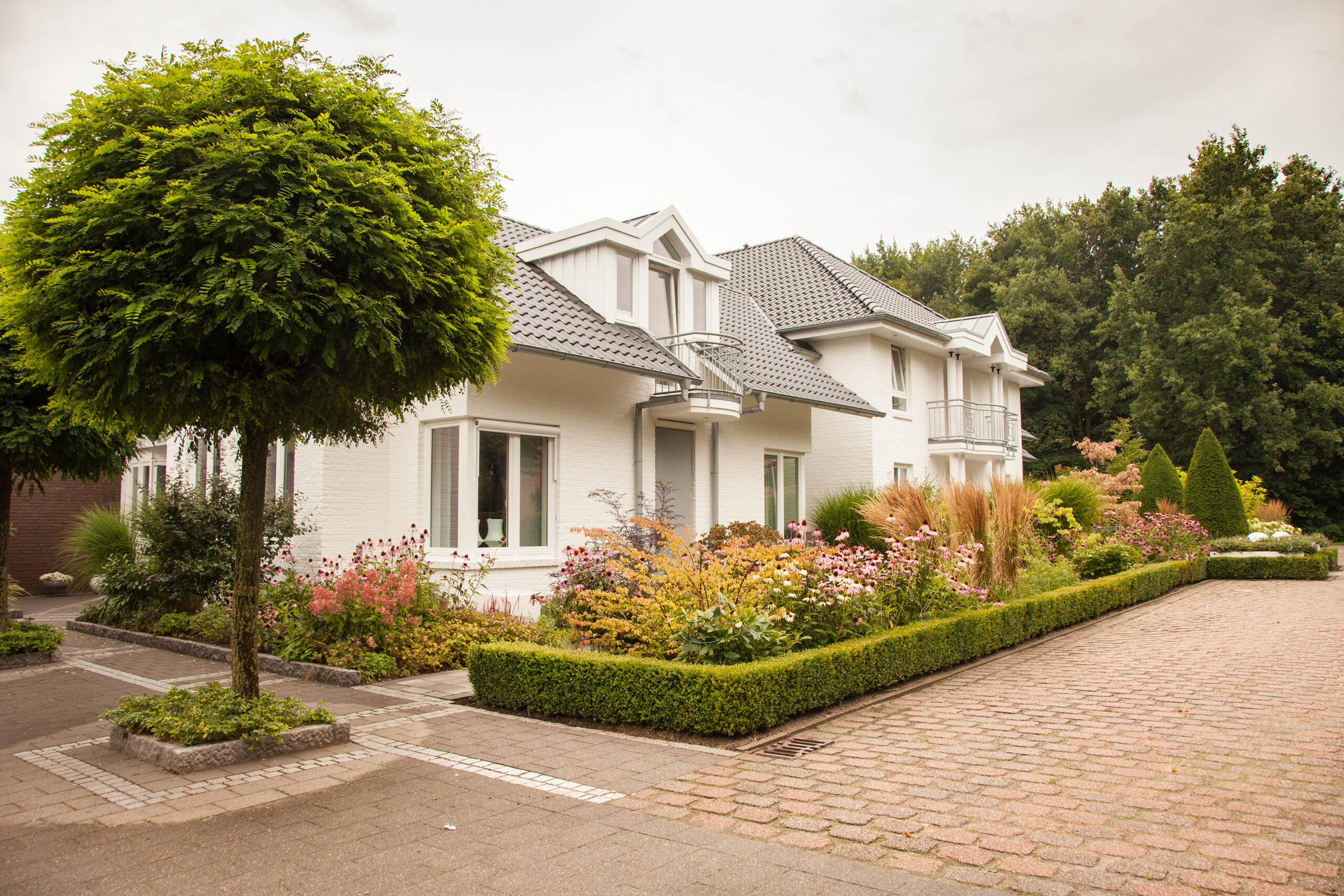 Modernes weißes zweistöckiges Haus mit grauem Dach, umgeben von einem üppigen, blühenden Garten.