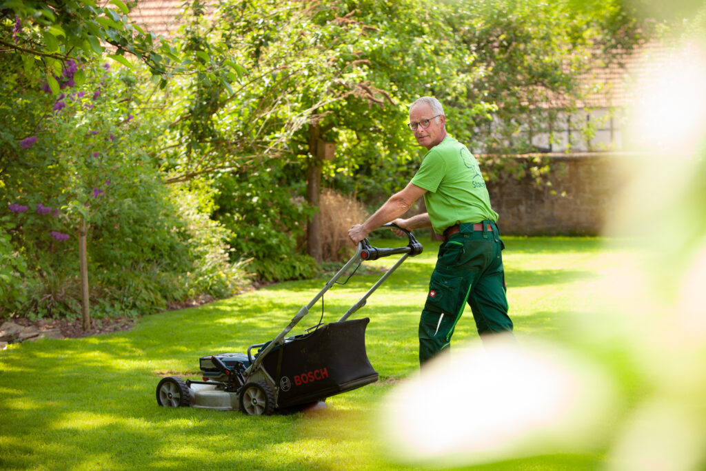Ein Mann in grüner Arbeitskleidung mäht mit einem Bosch-Rasenmäher einen gepflegten, grünen Rasen.