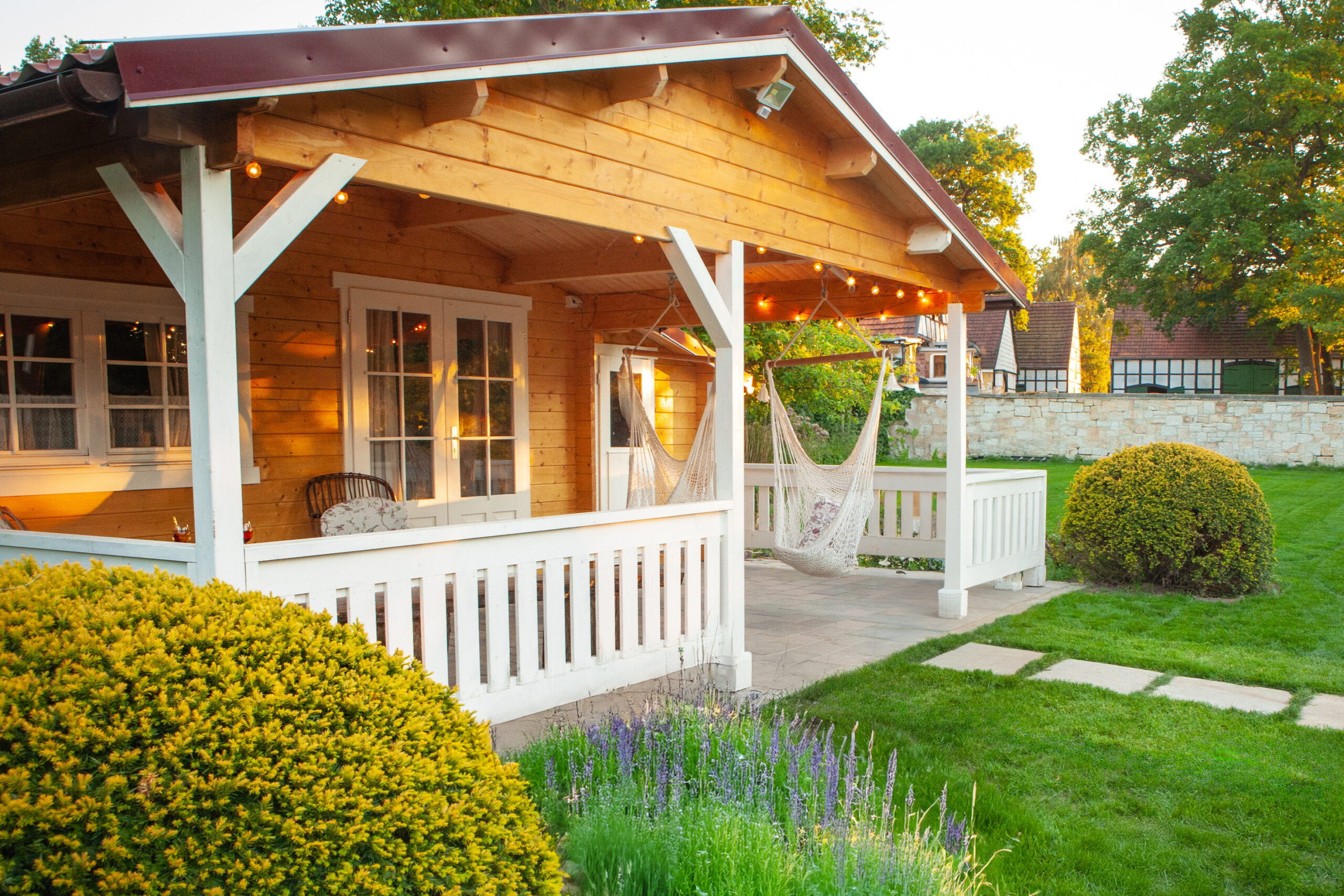 Gemütliche Veranda eines Holzhauses mit Hängesesseln, umgeben von gepflegtem Rasen und Büschen.