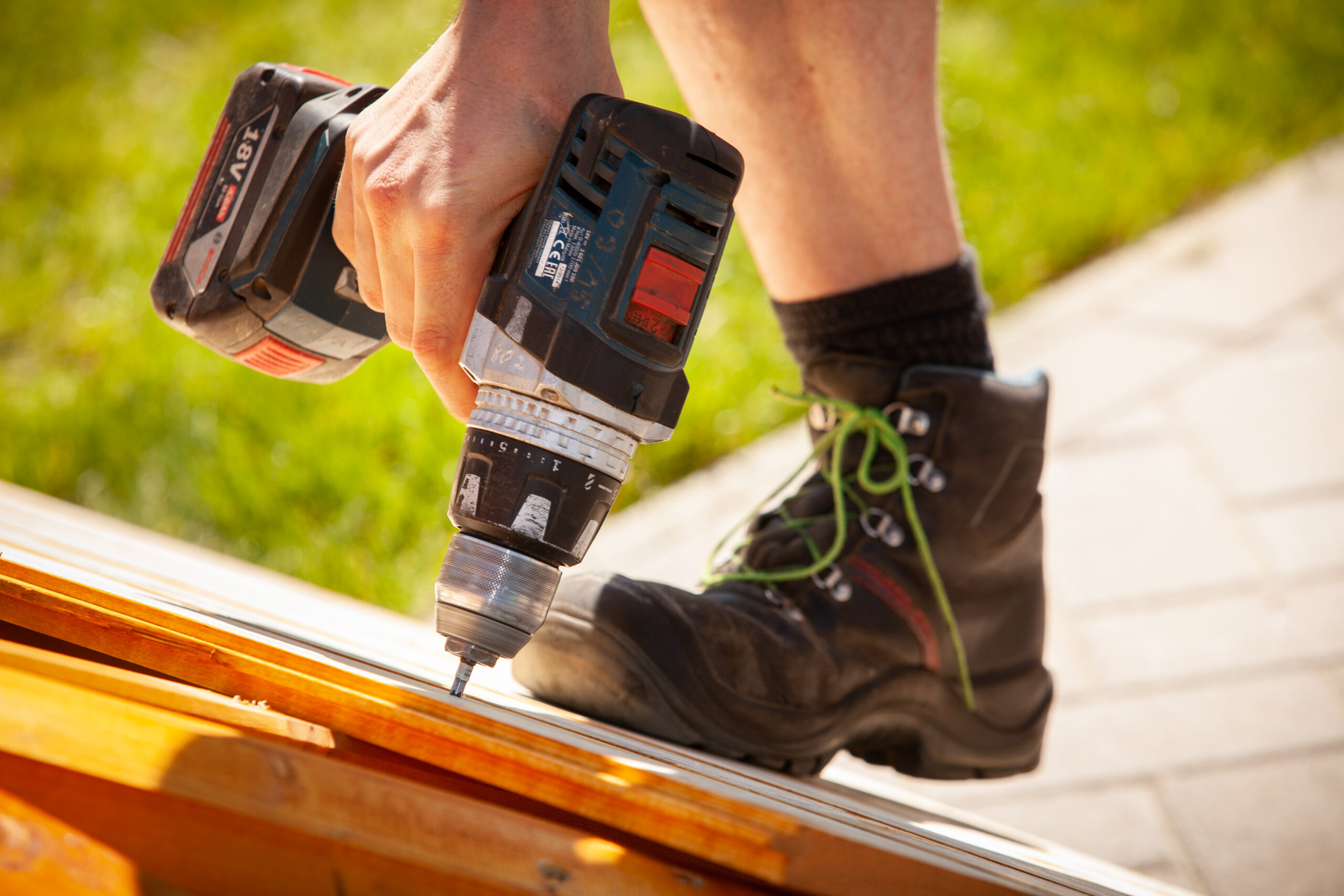 Person arbeitet mit Akkuschrauber an Holz, robustes Schuhwerk sichtbar, Outdoor-Umgebung.