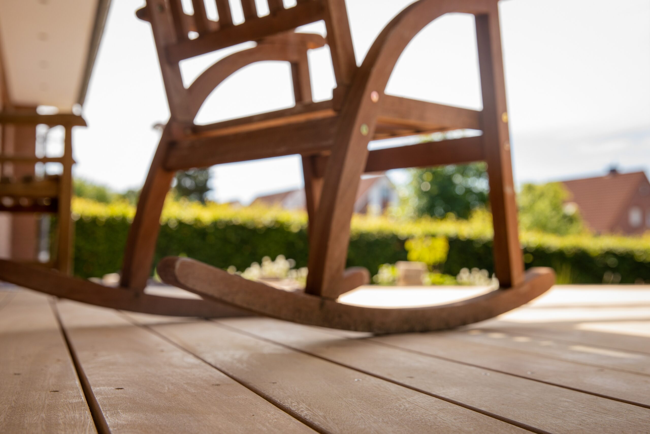 Ein hölzerner Schaukelstuhl steht still auf einer Terrasse mit Holzdielen, umgeben von grüner Natur.