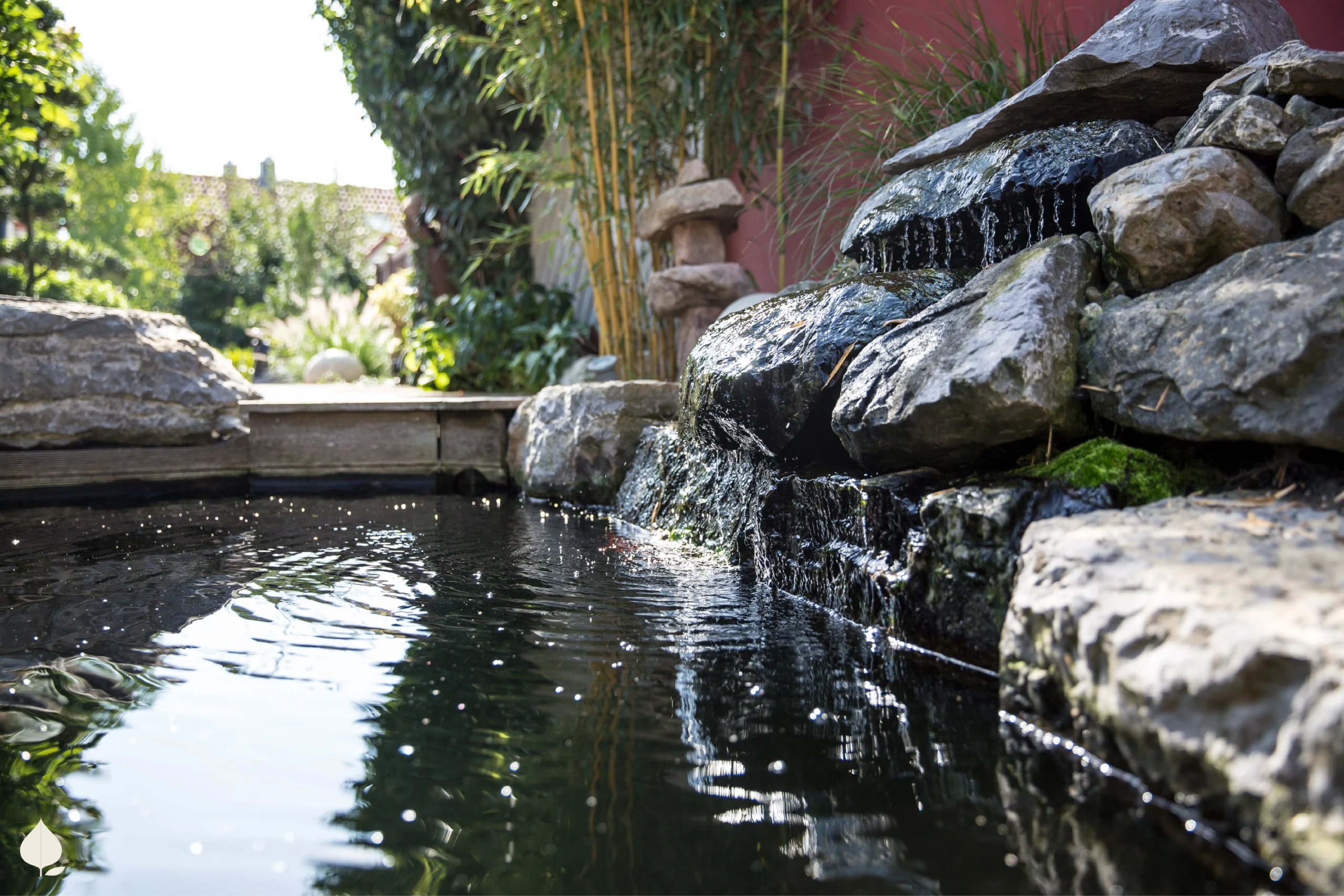 Ein ruhiger Gartenteich mit kleinem Wasserfall, umgeben von Pflanzen und Bambus, strahlt Frieden aus.