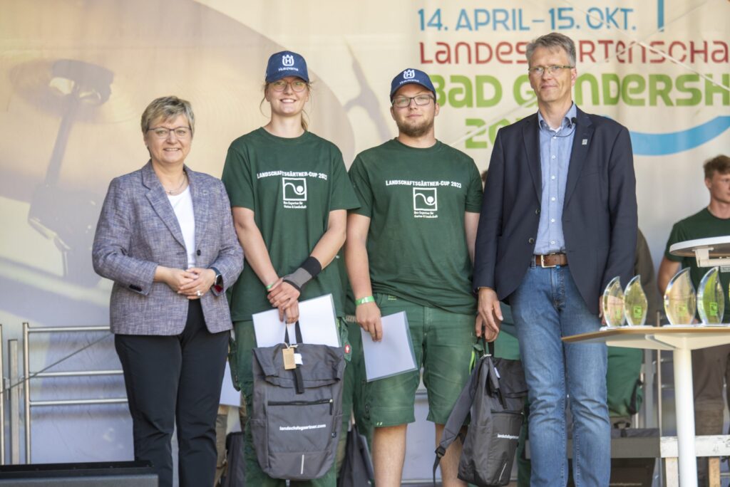 Vier Personen posieren auf einer Bühne; zwei in grüner Arbeitskleidung, zwei formell, mit Trophäen im Hintergrund.