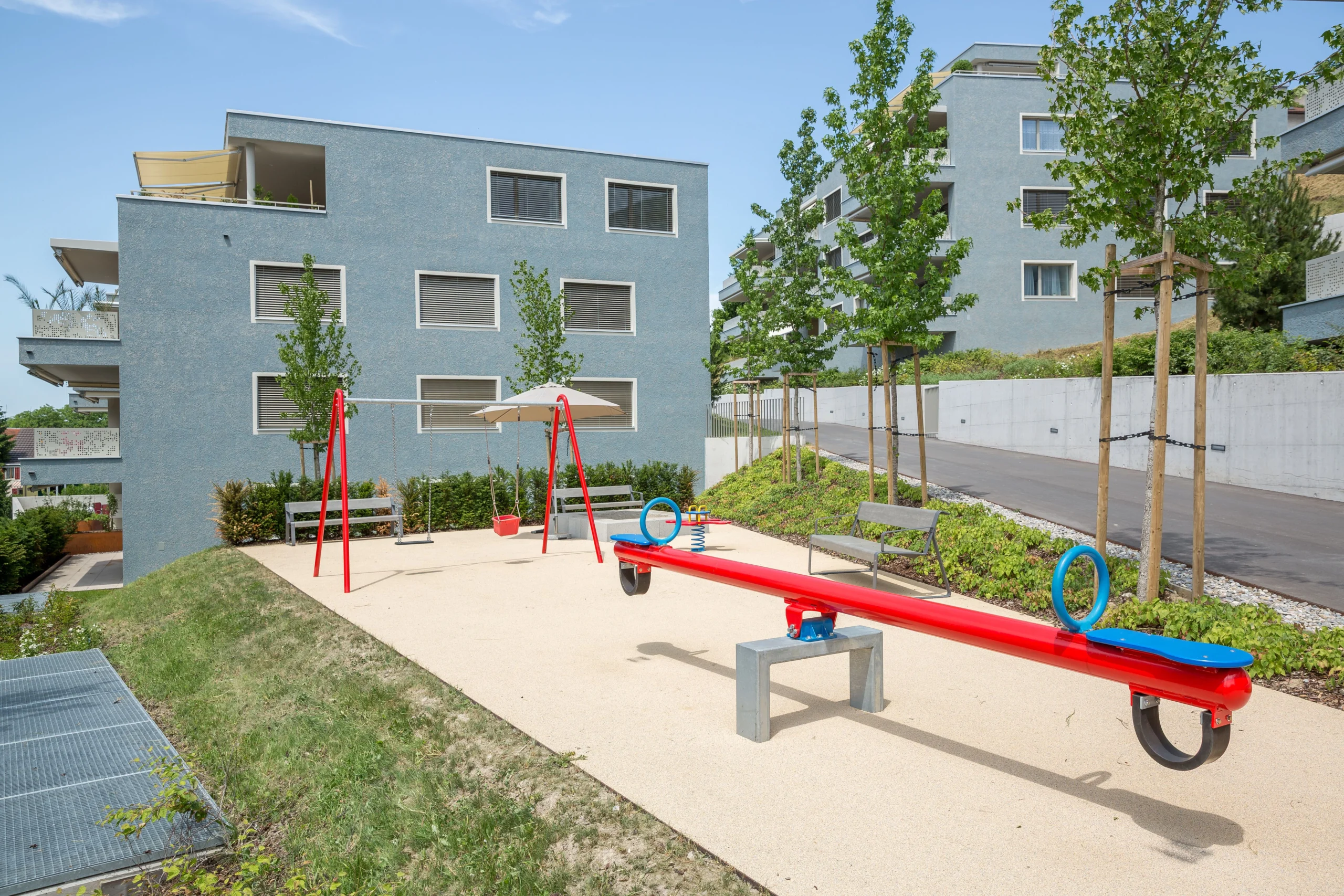 Moderner Spielplatz mit roten Spielgeräten und blauen Wohngebäuden im Hintergrund, umgeben von Grün.