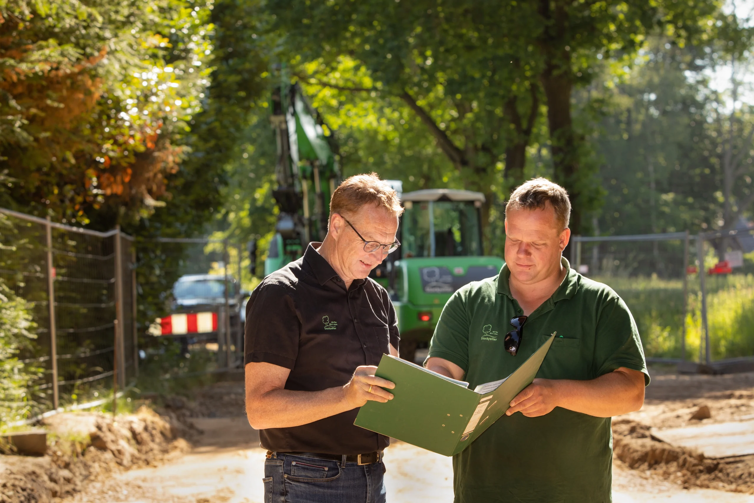 Zwei Männer in Arbeitskleidung besprechen Pläne auf einer Baustelle mit einem Bagger im Hintergrund.