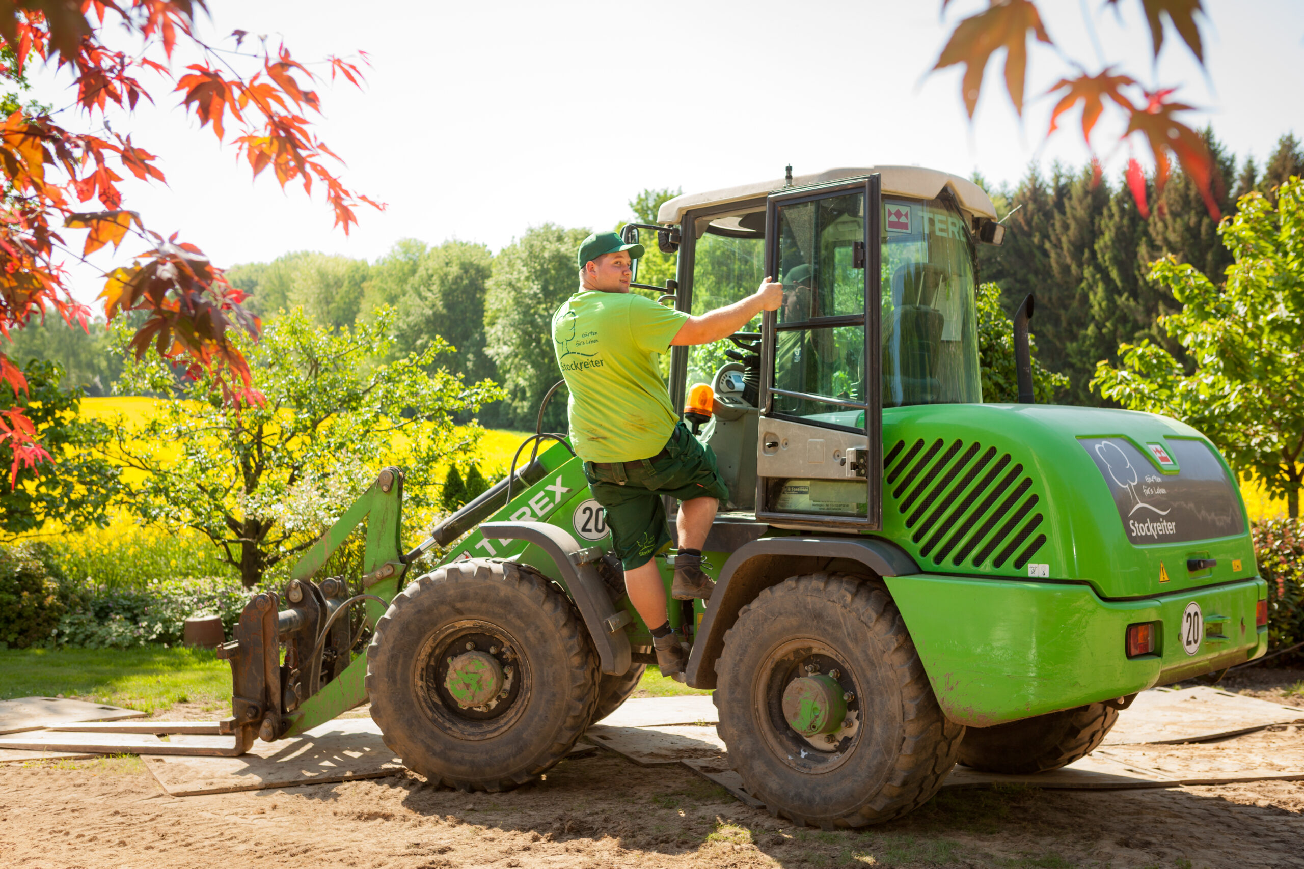 Ein Mann in grüner Kleidung steigt in einen grünen Bagger in einer ländlichen, blühenden Umgebung.