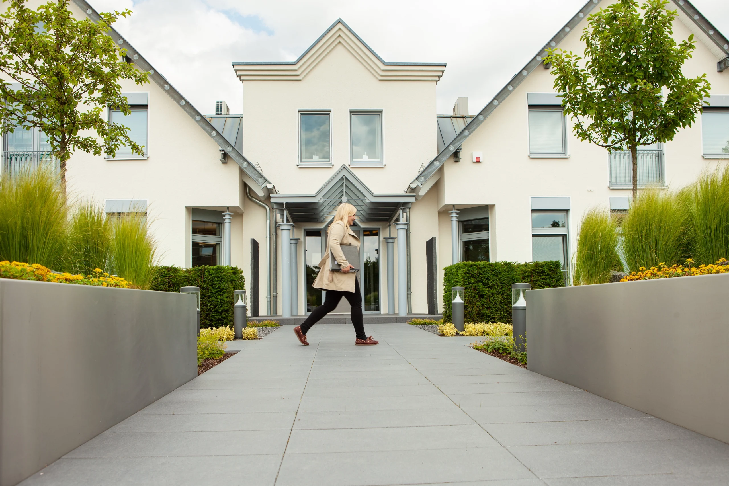 Moderne Gebäudefront mit großen Fenstern, gepflegtem Garten; Person geht zum Eingang.
