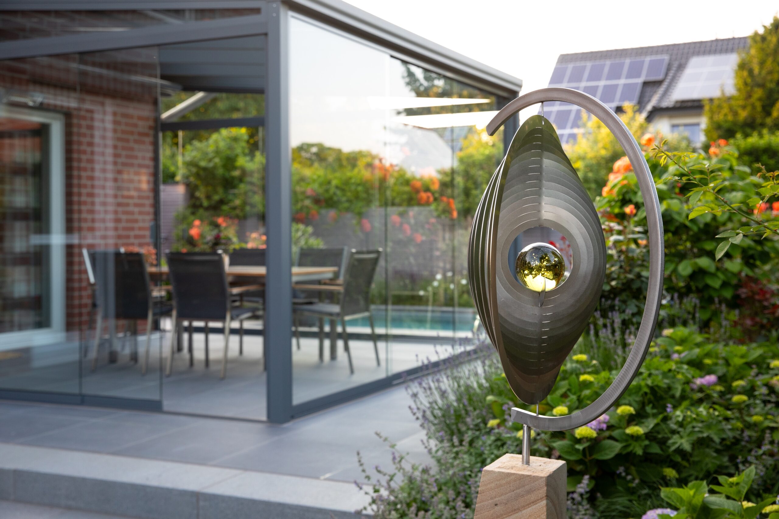 Moderne Gartenszene mit metallischer Skulptur und verglastem Pavillon in einem üppig grünen Umfeld.