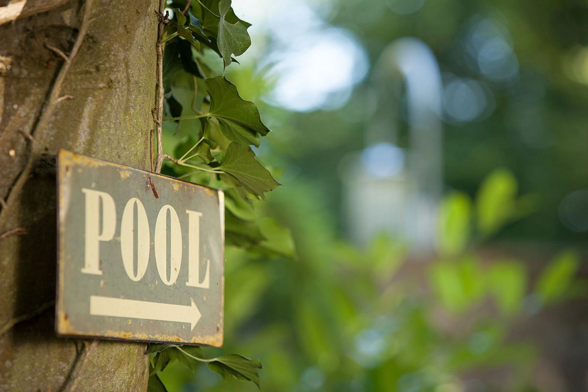 Ein Metallschild mit "POOL" und Pfeil zeigt an einem Baum befestigt den Weg zu einem Poolbereich.