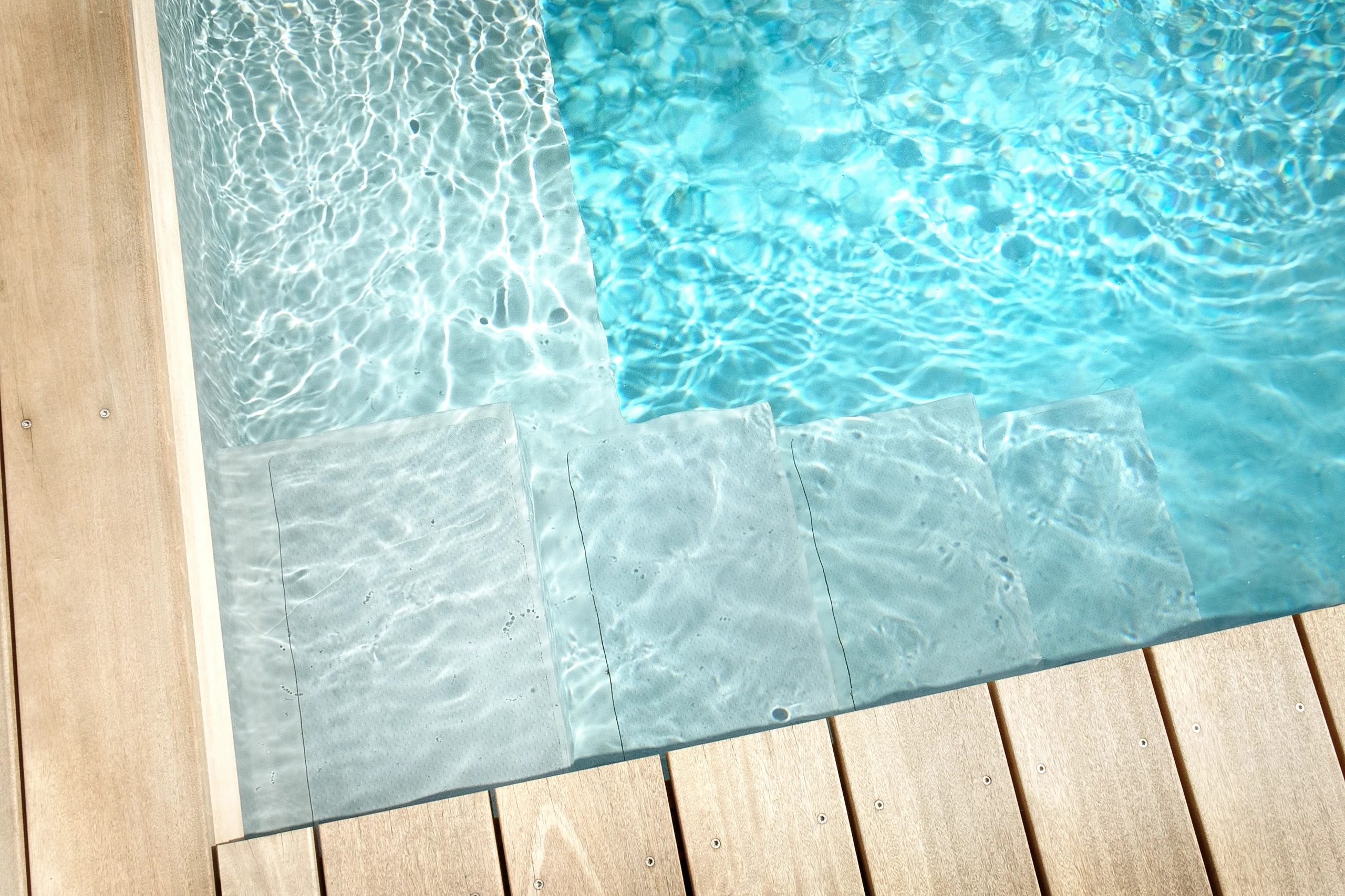 Ein ruhiger Pool mit blauen Wasser, einer grauen Treppe und einem hellbraunen Holzdeck. Keine Menschen.